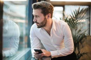 Un homme pensif avec un smartphone regardant par la fenêtre d'un bureau.
