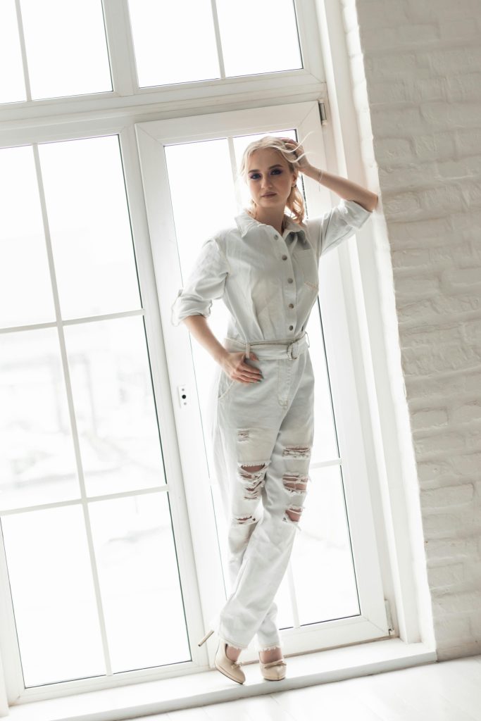 Stylish woman in a white jumpsuit poses by a large window, exuding modern fashion. Une femme élégante en combinaison blanche pose près d'une grande fenêtre, dégageant une mode moderne.