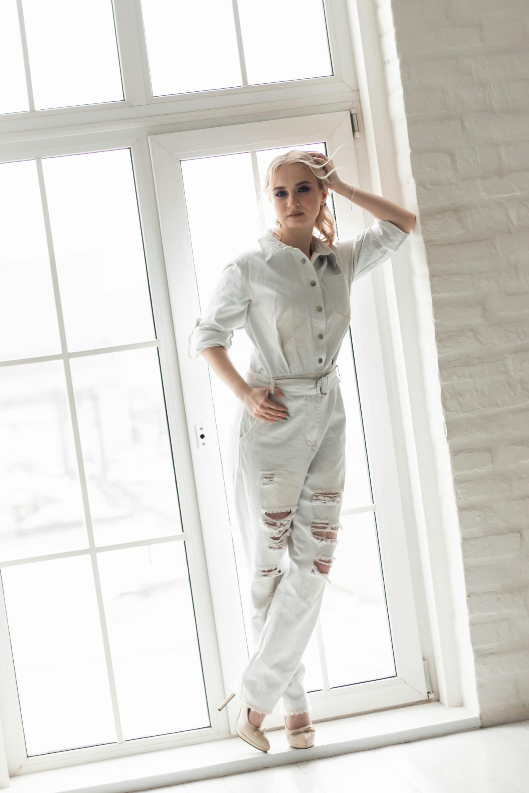 Stylish woman in a white jumpsuit poses by a large window, exuding modern fashion. Une femme élégante en combinaison blanche pose près d'une grande fenêtre, dégageant une mode moderne.
