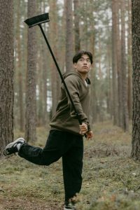 A young man in casual attire playfully swings a broomstick amidst a tranquil forest setting. Un jeune homme en tenue décontractée balance jouablement un balai au milieu d'un cadre forestier tranquille.