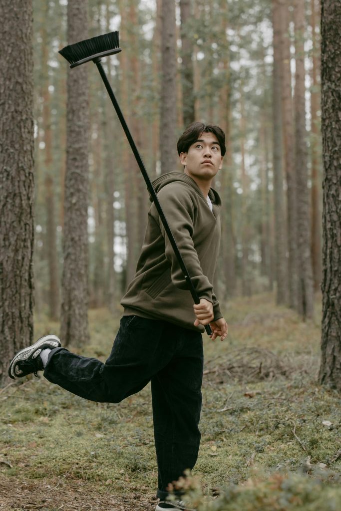 A young man in casual attire playfully swings a broomstick amidst a tranquil forest setting. Un jeune homme en tenue décontractée balance jouablement un balai au milieu d'un cadre forestier tranquille.