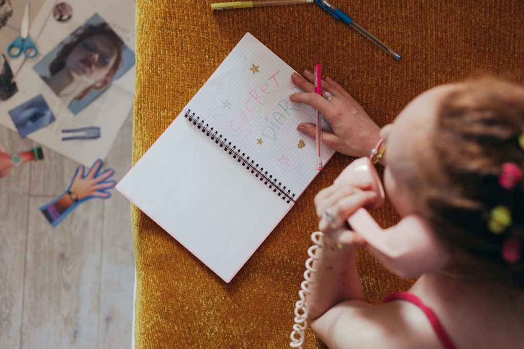 Woman writing in a diary while on a vintage pink phone, highlighting nostalgia and creativity. Une femme écrivant dans un journal intime tout en parlant sur un téléphone rose vintage, mettant en évidence la nostalgie et la créativité.