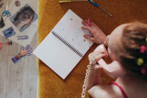 Woman writing in a diary while on a vintage pink phone, highlighting nostalgia and creativity. Une femme écrivant dans un journal intime tout en parlant sur un téléphone rose vintage, mettant en évidence la nostalgie et la créativité.