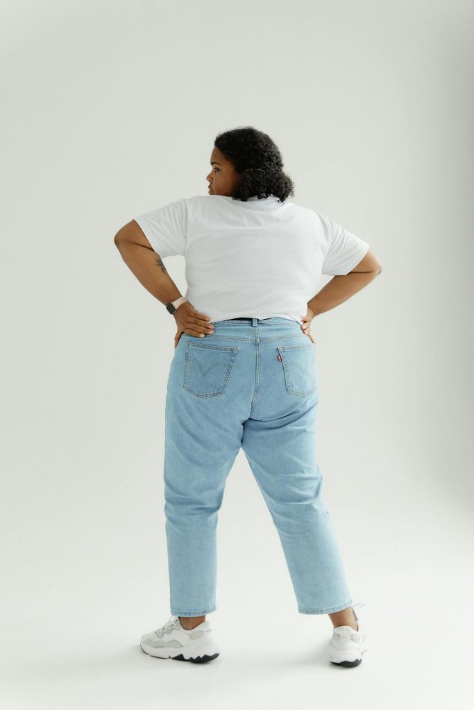 Vue arrière d'une femme confiante de grande taille en pantalon denim et chemise blanche debout sur fond blanc, embrassant la positivité corporelle.