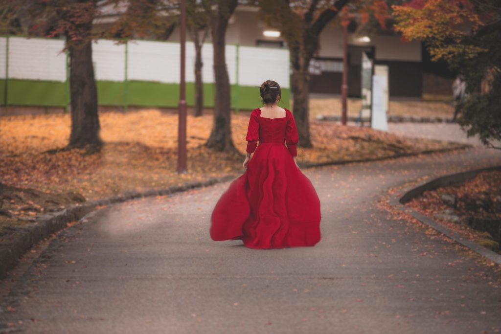 A woman in a red gown walks down a leafy autumn path, creating a romantic and elegant scene. Une femme en robe rouge marche le long d'un chemin automnal feuillu, créant une scène romantique et élégante.