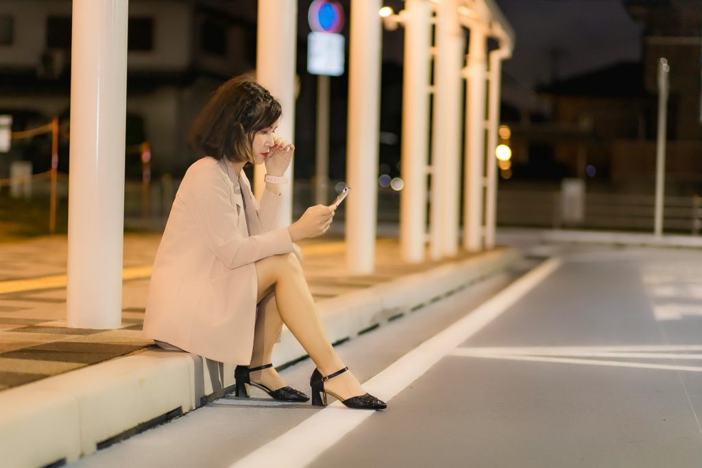 A woman sitting on a sidewalk using her smartphone at night under urban lights. Une femme assise sur un trottoir utilisant son smartphone la nuit sous des lumières urbaines.