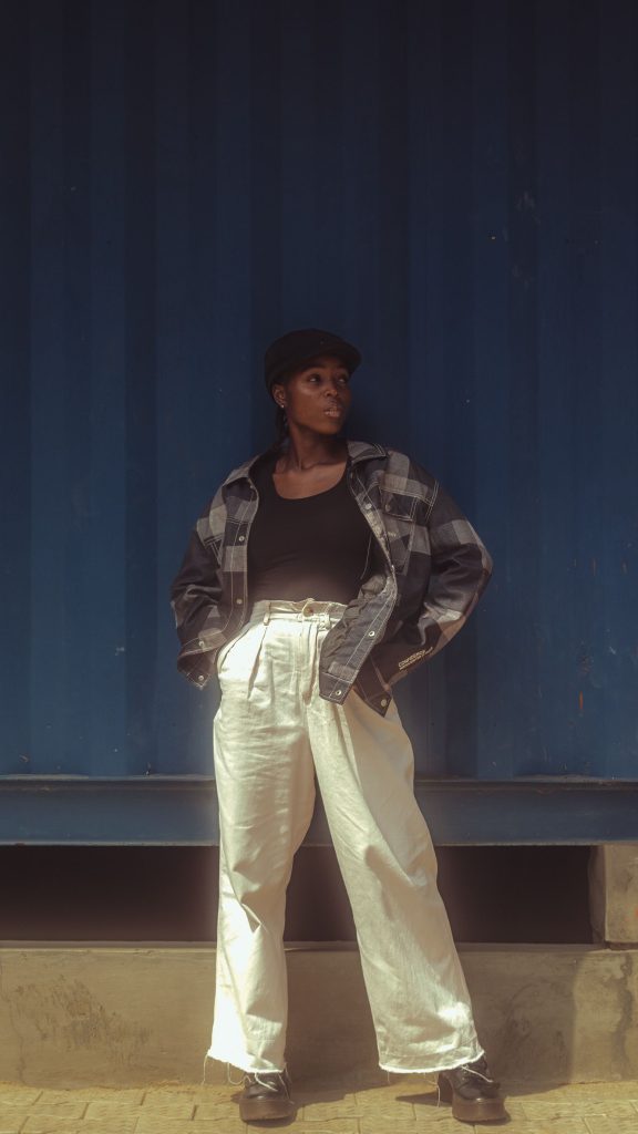 Woman in trendy outfit poses confidently by a blue wall, showcasing urban fashion. Femme en tenue tendance posant avec assurance devant un mur bleu, mettant en avant la mode urbaine.