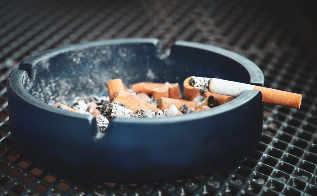 Close-up of cigarette butts and ashes in a blue ashtray with a smoldering cigarette, highlighting smoking habits. Gros plan sur des mégots de cigarettes et des cendres dans un cendrier bleu avec une cigarette qui fume, mettant en évidence les habitudes de tabagisme.